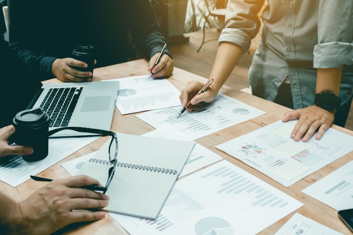 Several people in casual clothes with coffee and pens at a plywood table with charts and graphs on it.