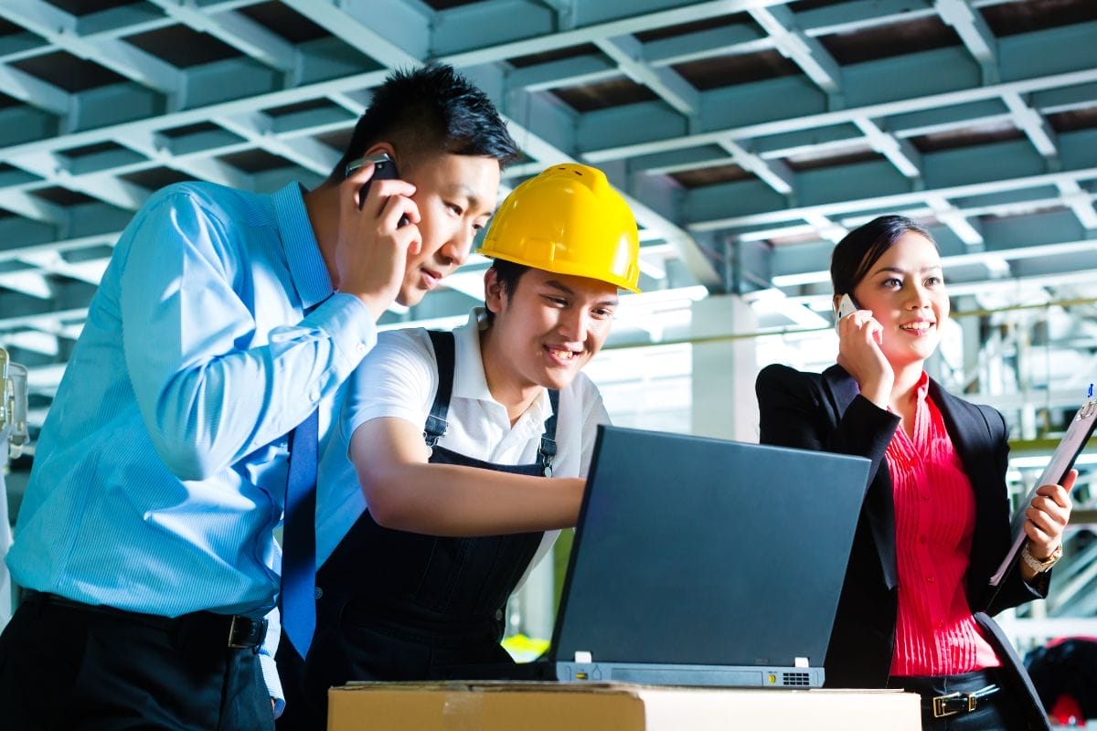 A woman with a notepad on a cell phone, plus a man on his cell phone, and a woman in a yellow hard hat, looking at a laptop screen.
