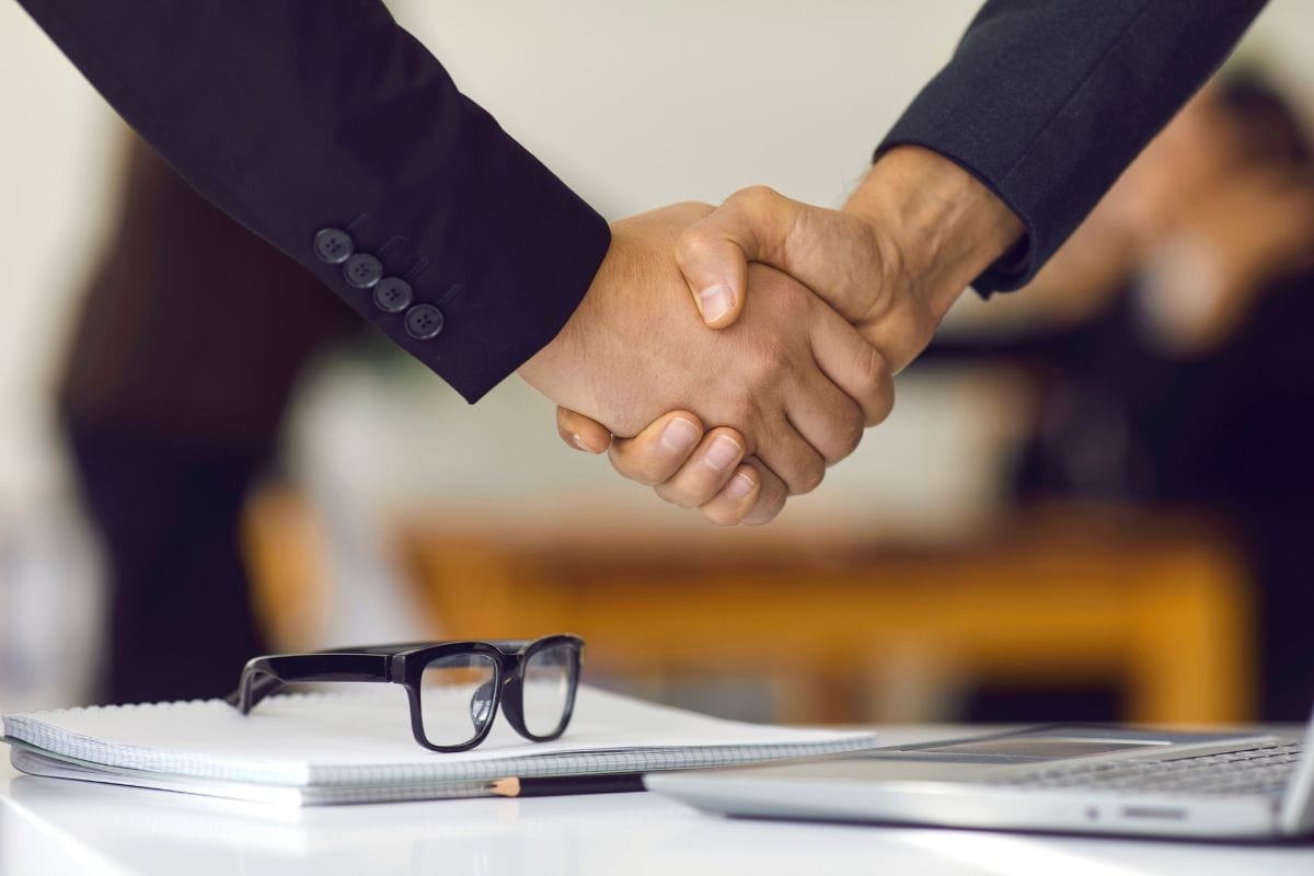 Men shaking hands and below are eyeglasses, a laptop and a notepad on a table.