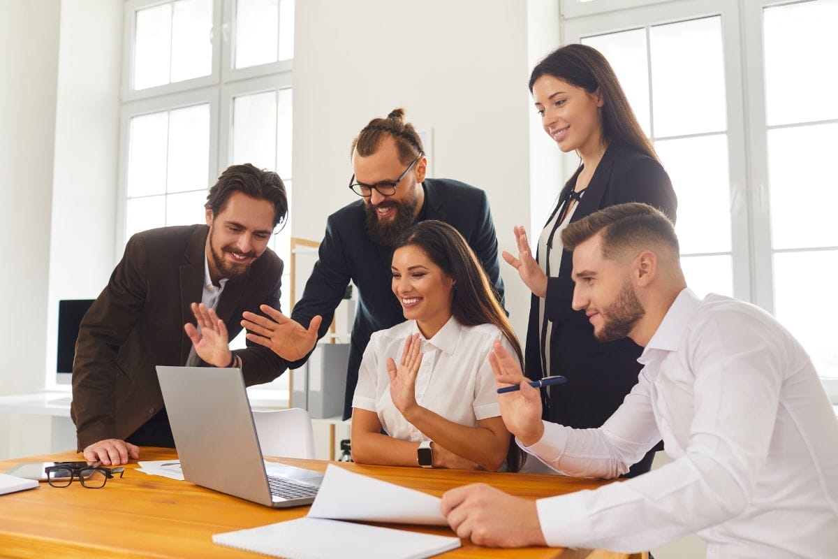 A group of 5 young professionals at a boardroom table waving to a Zoom caller on a laptop.